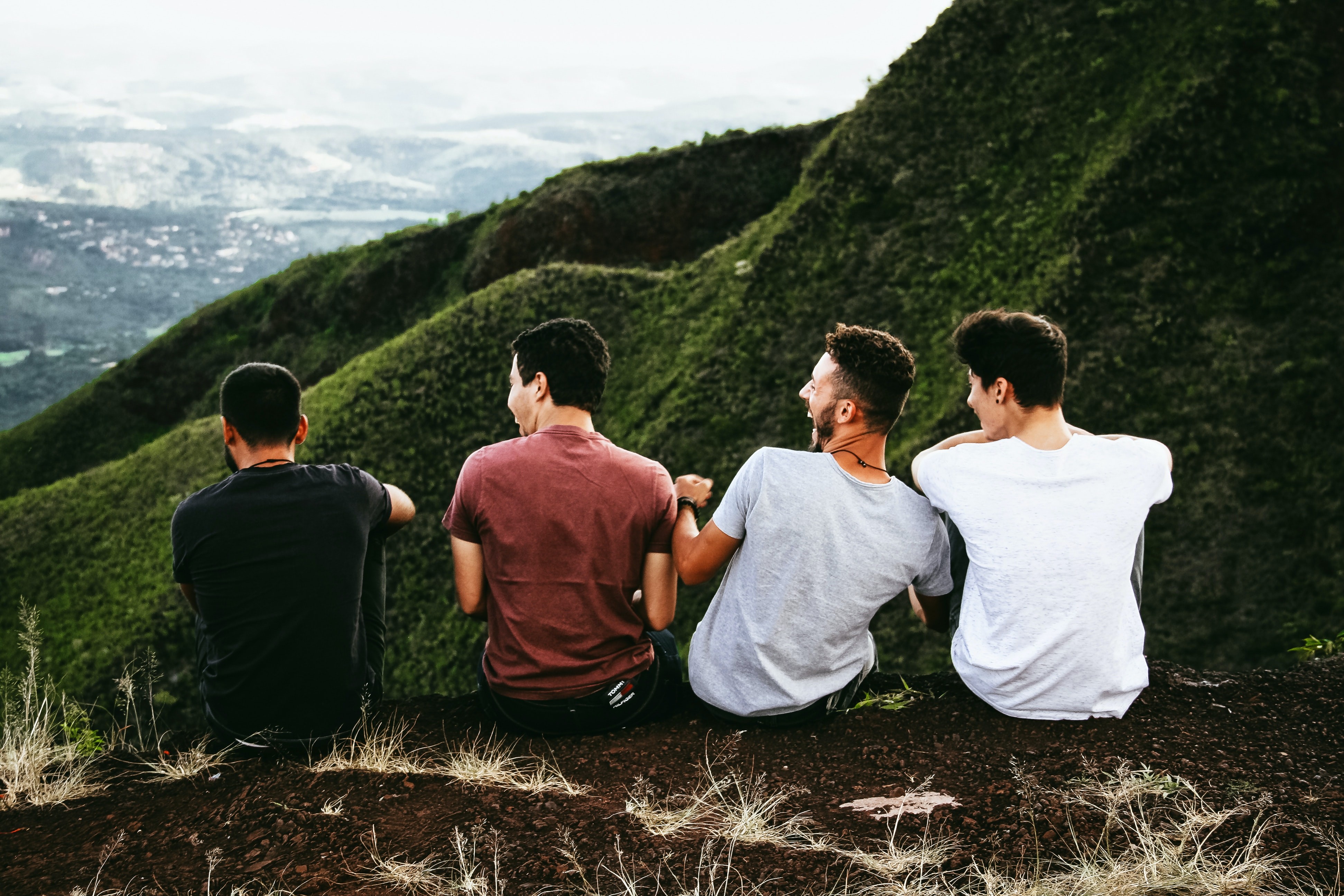 photo of four men sat on a hill, laughing, taken from behind them, looking over them at the view